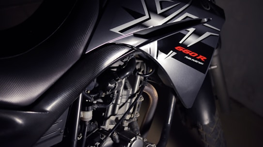 Close-up view of a motorcycle with a focus on its sleek black design featuring bold angular patterns and the label '660R Fuel Injection' highlighted in red. The image emphasizes the mechanical components, showcasing the engine and part of the exhaust.