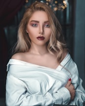 A woman with blonde wavy hair, wearing an off-shoulder white shirt, is looking directly at the camera with a serious expression. Her makeup is bold with dark red lipstick and well-defined eyes. There are blurred lights in the background, giving a bokeh effect.