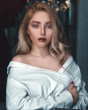 A woman with blonde wavy hair, wearing an off-shoulder white shirt, is looking directly at the camera with a serious expression. Her makeup is bold with dark red lipstick and well-defined eyes. There are blurred lights in the background, giving a bokeh effect.