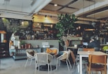 A cozy cafe with a contemporary design featuring various seating arrangements including wicker chairs, wooden chairs, and tables. The counter displays potted plants, glassware, and coffee equipment. The blackboard behind the counter has handwritten menu options. A person is sitting at a table working on a laptop. The environment includes warm lighting with hanging light fixtures and a large indoor plant providing a touch of greenery.