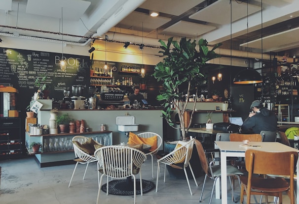 A cozy cafe with a contemporary design featuring various seating arrangements including wicker chairs, wooden chairs, and tables. The counter displays potted plants, glassware, and coffee equipment. The blackboard behind the counter has handwritten menu options. A person is sitting at a table working on a laptop. The environment includes warm lighting with hanging light fixtures and a large indoor plant providing a touch of greenery.