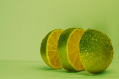A fresh lime cut into three segments is arranged in front of a matching green background. The fruit is positioned at an angle, highlighting its juicy, vibrant interiors.