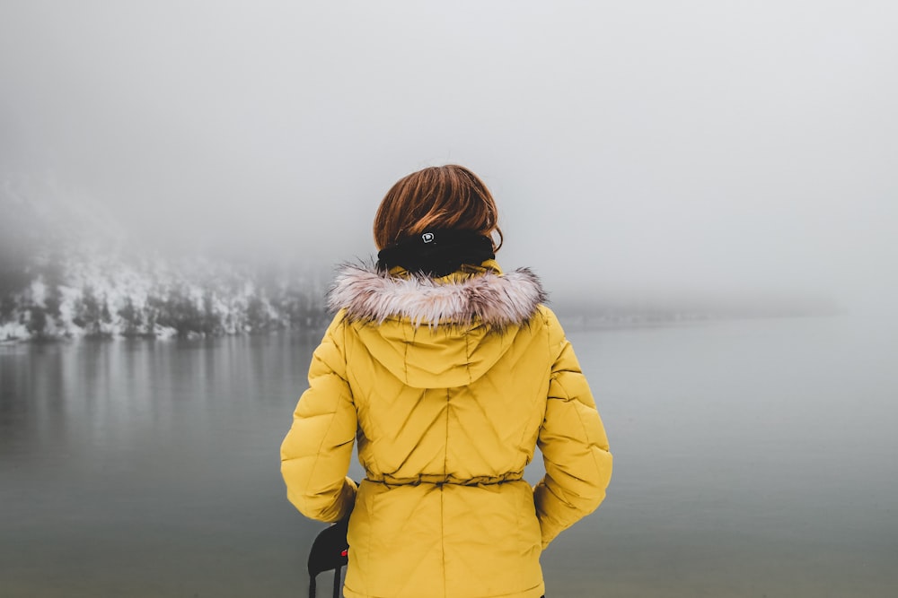 Mujer con abrigo amarillo mirando el cuerpo de agua cubierto de niebla