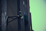 A surveillance camera mounted on the corner of a building, attached to a metal support structure. The building wall appears weathered and the sky in the background is a muted teal color.