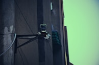A surveillance camera mounted on the corner of a building, attached to a metal support structure. The building wall appears weathered and the sky in the background is a muted teal color.