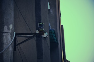 A surveillance camera mounted on the corner of a building, attached to a metal support structure. The building wall appears weathered and the sky in the background is a muted teal color.