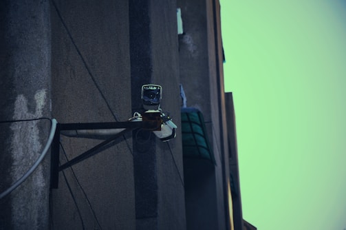 A surveillance camera mounted on the corner of a building, attached to a metal support structure. The building wall appears weathered and the sky in the background is a muted teal color.