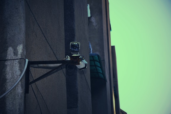 A surveillance camera mounted on the corner of a building, attached to a metal support structure. The building wall appears weathered and the sky in the background is a muted teal color.