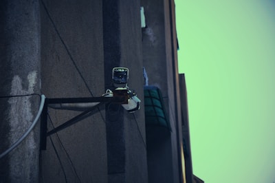 A surveillance camera mounted on the corner of a building, attached to a metal support structure. The building wall appears weathered and the sky in the background is a muted teal color.