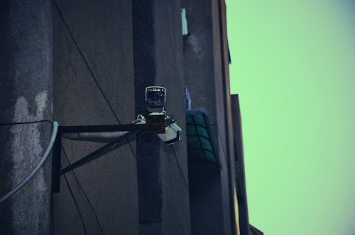 A surveillance camera mounted on the corner of a building, attached to a metal support structure. The building wall appears weathered and the sky in the background is a muted teal color.