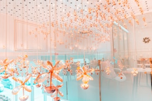 An elegant room with a ceiling adorned with numerous hanging golden floral ornaments. The space has a modern, luxurious design with glass walls and soft lighting. Chairs and tables can be seen in the background, contributing to a sophisticated and inviting atmosphere.