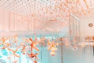 An elegant room with a ceiling adorned with numerous hanging golden floral ornaments. The space has a modern, luxurious design with glass walls and soft lighting. Chairs and tables can be seen in the background, contributing to a sophisticated and inviting atmosphere.
