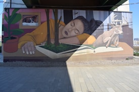 A mural on a wall depicting a woman with her eyes closed, resting her head on a large open book, which has plants growing from it. A deer stands beside her, and a window shows a dog's face peering out. There is a potted plant on the left side of the mural. The image is partially shadowed by the structure above.