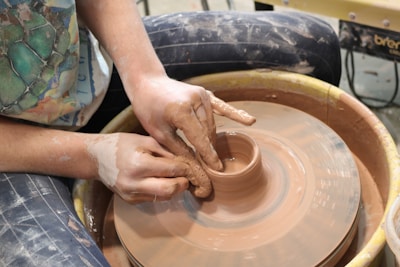 Hands are skillfully shaping clay on a spinning pottery wheel. The focus is on the creation of a clay pot, with the person's hands covered in moist clay, emphasizing the tactile nature of pottery.