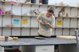 A person is intently examining a pottery piece in a studio. The background features shelves filled with various pottery tools and materials. The person is wearing a casual shirt and has their hair tied up.