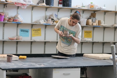 A person is intently examining a pottery piece in a studio. The background features shelves filled with various pottery tools and materials. The person is wearing a casual shirt and has their hair tied up.