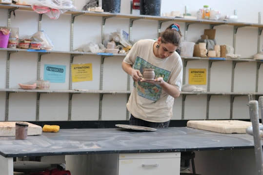 A person is intently examining a pottery piece in a studio. The background features shelves filled with various pottery tools and materials. The person is wearing a casual shirt and has their hair tied up.
