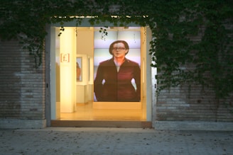 The image depicts the entrance of a building with a large glass door, surrounded by brick walls and climbing ivy. Inside, there is a prominent digital display showing an image of a person, adding a modern touch to the otherwise classic exterior. The interior is softly lit, creating an inviting atmosphere.