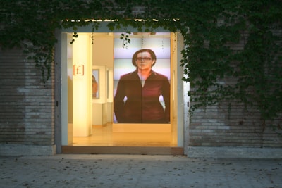 The image depicts the entrance of a building with a large glass door, surrounded by brick walls and climbing ivy. Inside, there is a prominent digital display showing an image of a person, adding a modern touch to the otherwise classic exterior. The interior is softly lit, creating an inviting atmosphere.