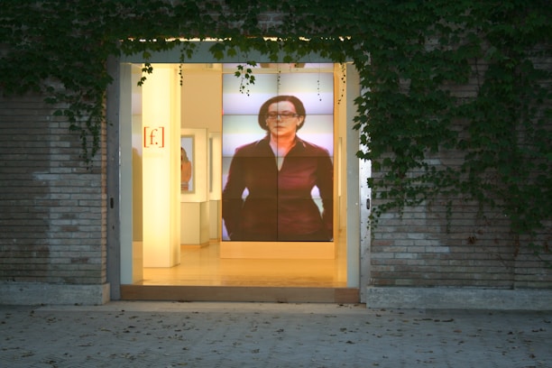 The image depicts the entrance of a building with a large glass door, surrounded by brick walls and climbing ivy. Inside, there is a prominent digital display showing an image of a person, adding a modern touch to the otherwise classic exterior. The interior is softly lit, creating an inviting atmosphere.