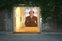 The image depicts the entrance of a building with a large glass door, surrounded by brick walls and climbing ivy. Inside, there is a prominent digital display showing an image of a person, adding a modern touch to the otherwise classic exterior. The interior is softly lit, creating an inviting atmosphere.