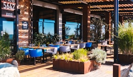 A stylish outdoor seating area of a restaurant with wooden decking and modern furniture. Tables are set with glasses, and the area is surrounded by lush greenery in planters. The building's facade is made of brick and glass, with a sign displaying the number 5642.