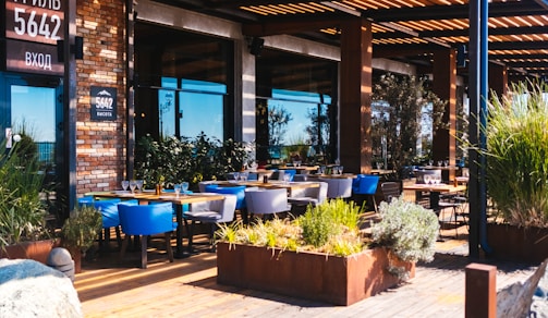 A stylish outdoor seating area of a restaurant with wooden decking and modern furniture. Tables are set with glasses, and the area is surrounded by lush greenery in planters. The building's facade is made of brick and glass, with a sign displaying the number 5642.