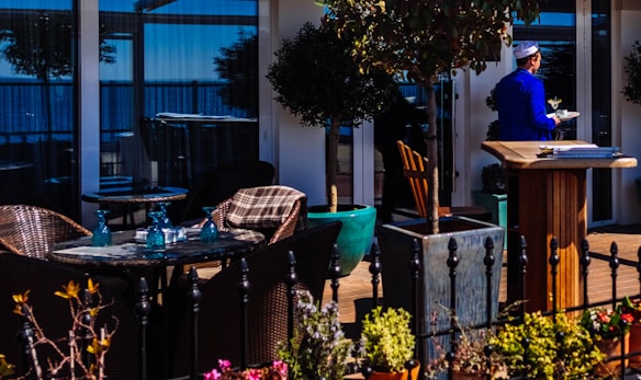 A cozy outdoor restaurant setting with wicker tables and chairs, adorned with blue glass bottles. Potted plants add greenery to the space, while a man in a blue uniform and hat carries a tray with drinks through the area. The background features large glass windows reflecting the blue sky and trees.