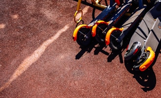 Several electric scooters with bright orange front wheels are neatly lined up on a reddish-brown textured ground. The sunlight casts distinct shadows, emphasizing the vibrant colors.
