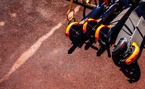 Several electric scooters with bright orange front wheels are neatly lined up on a reddish-brown textured ground. The sunlight casts distinct shadows, emphasizing the vibrant colors.