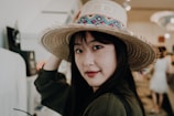 A woman with long dark hair is wearing a large straw hat adorned with colorful patterns. She is indoors, possibly in a shop or a market, and looks over her shoulder with a slight smile. The background is slightly blurred, featuring people and market stalls, suggesting a bustling environment.