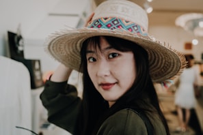 A woman with long dark hair is wearing a large straw hat adorned with colorful patterns. She is indoors, possibly in a shop or a market, and looks over her shoulder with a slight smile. The background is slightly blurred, featuring people and market stalls, suggesting a bustling environment.