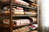 A wooden shelf filled with neatly folded, colorful quilts arranged in stacks. The quilts display a variety of patterns, including geometric shapes and floral designs. A sheer curtain with intricate floral motifs hangs beside the shelf, filtering soft light into the space.