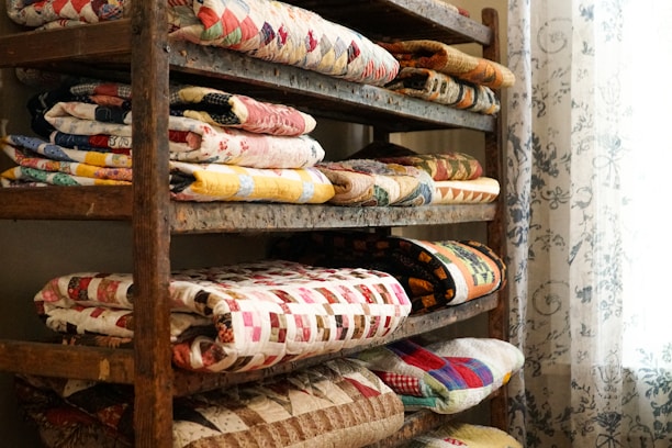 A wooden shelf filled with neatly folded, colorful quilts arranged in stacks. The quilts display a variety of patterns, including geometric shapes and floral designs. A sheer curtain with intricate floral motifs hangs beside the shelf, filtering soft light into the space.