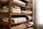 A wooden shelf filled with neatly folded, colorful quilts arranged in stacks. The quilts display a variety of patterns, including geometric shapes and floral designs. A sheer curtain with intricate floral motifs hangs beside the shelf, filtering soft light into the space.
