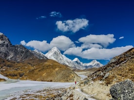 A vast mountainous landscape with snow-capped peaks under a clear blue sky. The scene is dominated by rugged terrain, rocky slopes, and a small frozen river winding through the valley. A solitary hiker, dressed in red, can be seen walking along a trail, emphasizing the scale and remoteness of the environment.