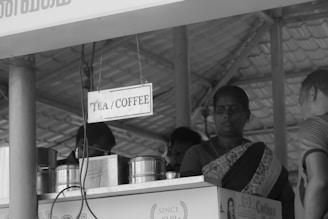 A black and white image features a small stall or kiosk with a sign labeled 'Tea/Coffee'. There are several metal containers on the counter, and a woman wearing a traditional outfit is standing next to them. The roof and structure of the kiosk are made of corrugated metal.