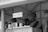 A black and white image features a small stall or kiosk with a sign labeled 'Tea/Coffee'. There are several metal containers on the counter, and a woman wearing a traditional outfit is standing next to them. The roof and structure of the kiosk are made of corrugated metal.