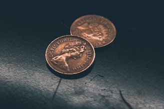 Two vintage copper-toned coins are placed on a dark, textured surface. The coins display detailed inscriptions and profiles, indicating they are of British origin, with one slightly overlapping the other.