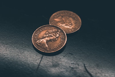 Two vintage copper-toned coins are placed on a dark, textured surface. The coins display detailed inscriptions and profiles, indicating they are of British origin, with one slightly overlapping the other.