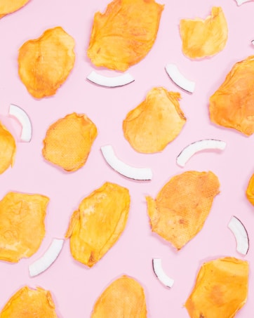 Bright yellow dried mango slices and white coconut strips are scattered randomly on a pastel pink background.