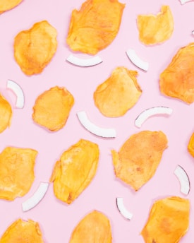 Bright yellow dried mango slices and white coconut strips are scattered randomly on a pastel pink background.