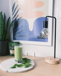 A glass of green smoothie sits on a round marble plate accompanied by fresh spinach leaves. In the background, there is an abstract pastel-colored artwork with shapes resembling leaves and waves. A modern lamp with a clear bulb is situated to the right, its base made of light wood.