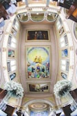 The image captures a detailed view of a church interior with a fisheye lens effect. The ceiling is adorned with intricate paintings depicting religious figures and scenes. The surrounding architecture includes arches and decorative moldings. White floral arrangements are visible on both sides, suggesting a ceremonial setting.