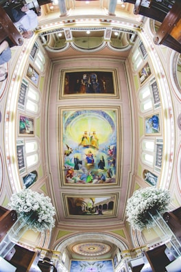 The image captures a detailed view of a church interior with a fisheye lens effect. The ceiling is adorned with intricate paintings depicting religious figures and scenes. The surrounding architecture includes arches and decorative moldings. White floral arrangements are visible on both sides, suggesting a ceremonial setting.