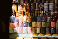 A variety of colorful pigment powders are displayed in plastic bags, stacked neatly in rows. Above them, various cans and bottles of paint, varnish, and other industrial products are organized. The setup appears to be from a market or shop selling art and construction materials.