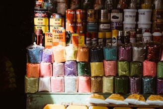 A variety of colorful pigment powders are displayed in plastic bags, stacked neatly in rows. Above them, various cans and bottles of paint, varnish, and other industrial products are organized. The setup appears to be from a market or shop selling art and construction materials.