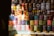 A variety of colorful pigment powders are displayed in plastic bags, stacked neatly in rows. Above them, various cans and bottles of paint, varnish, and other industrial products are organized. The setup appears to be from a market or shop selling art and construction materials.