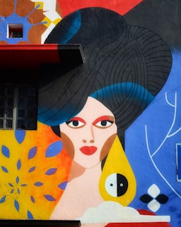 A vibrant mural featuring a stylized, abstract portrait of a woman with geometric shapes and bold colors. Her large, dark hair is intricately detailed, and her features are exaggerated with bright red lips and large eyes. Surrounding her are colorful patterns including blue leaves and an abstract sunburst.