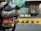 A motorcycle is carrying large, colorful woven bags filled with goods and small bouquets of greens. The background features a vibrant mural on a wall with the words 'LOVE DELHI' in bold white letters. There is a barred window next to the mural and a black and yellow striped curb. The scene is illuminated by sunlight, giving it a lively urban street vibe.